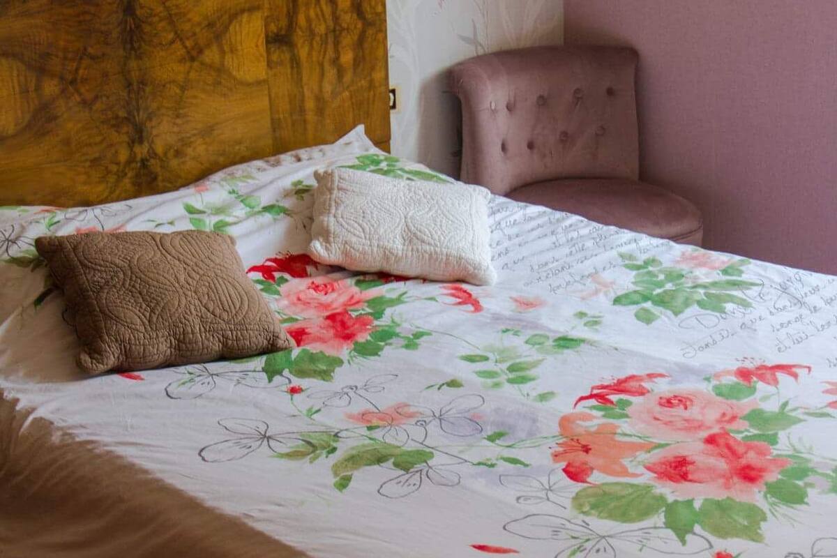 A neatly made bed with floral bedding, a wooden headboard, and a small armchair in a room with patterned wallpaper.