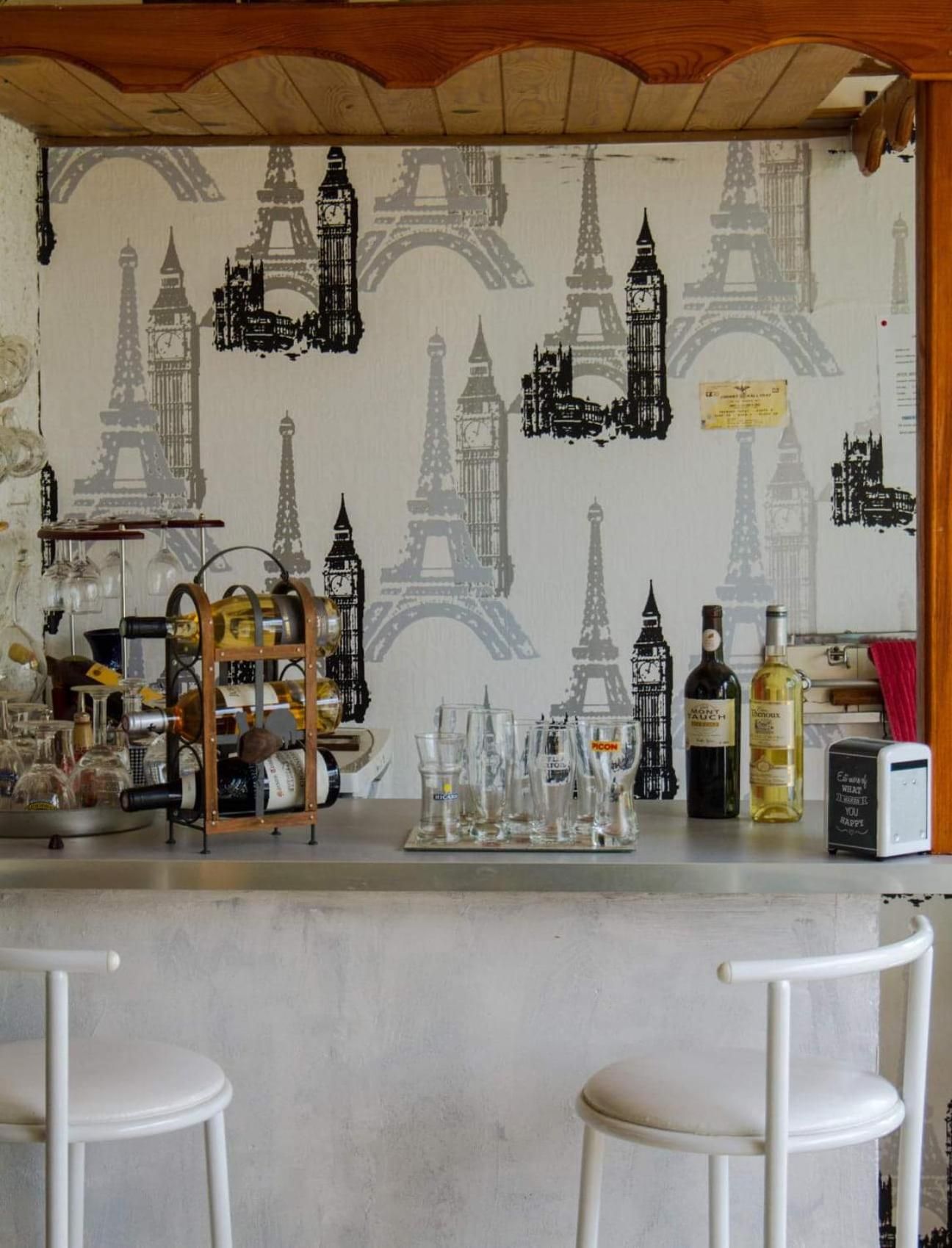A bar area with a Paris-themed wall, glasses, bottles, and a small refrigerator.