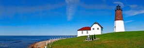 Un faro con techo rojo y paredes blancas, un edificio blanco con techo rojo, y un área verde con un banco.