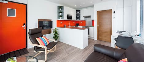 A modern living room with a sofa, a glass coffee table, a TV, and a kitchen area with white cabinets and an orange accent wall.