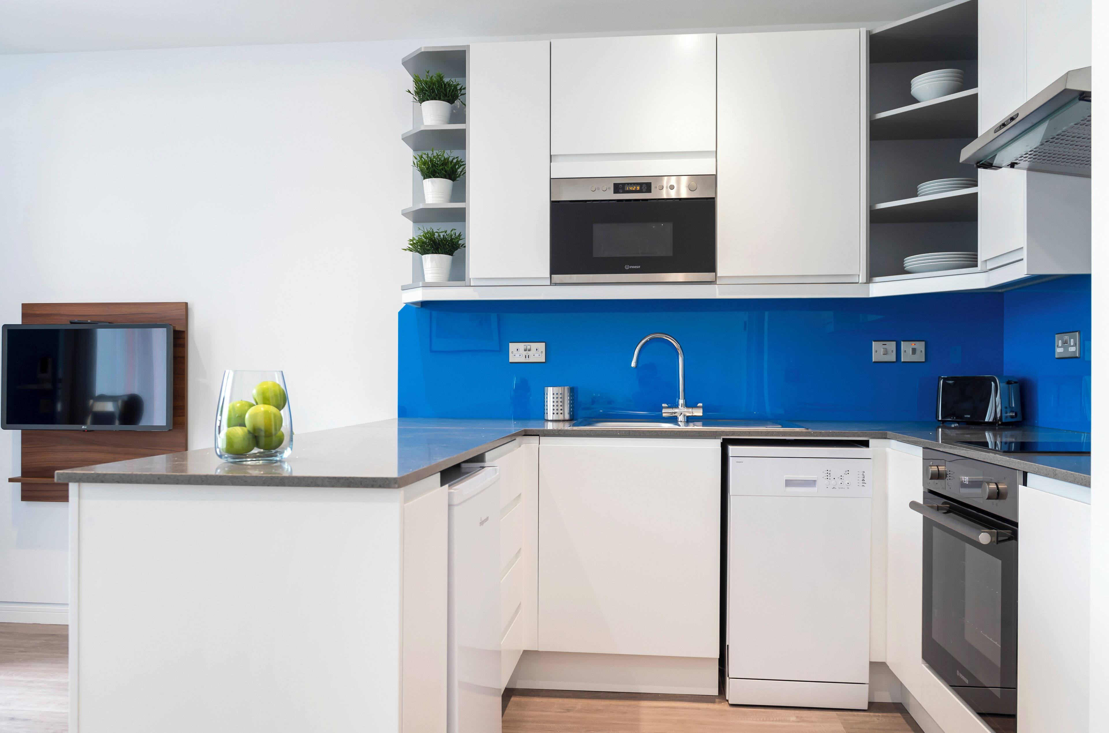 Uma cozinha moderna com armários brancos, painel de fundo azul, forno embutido e máquina de lavar louça.