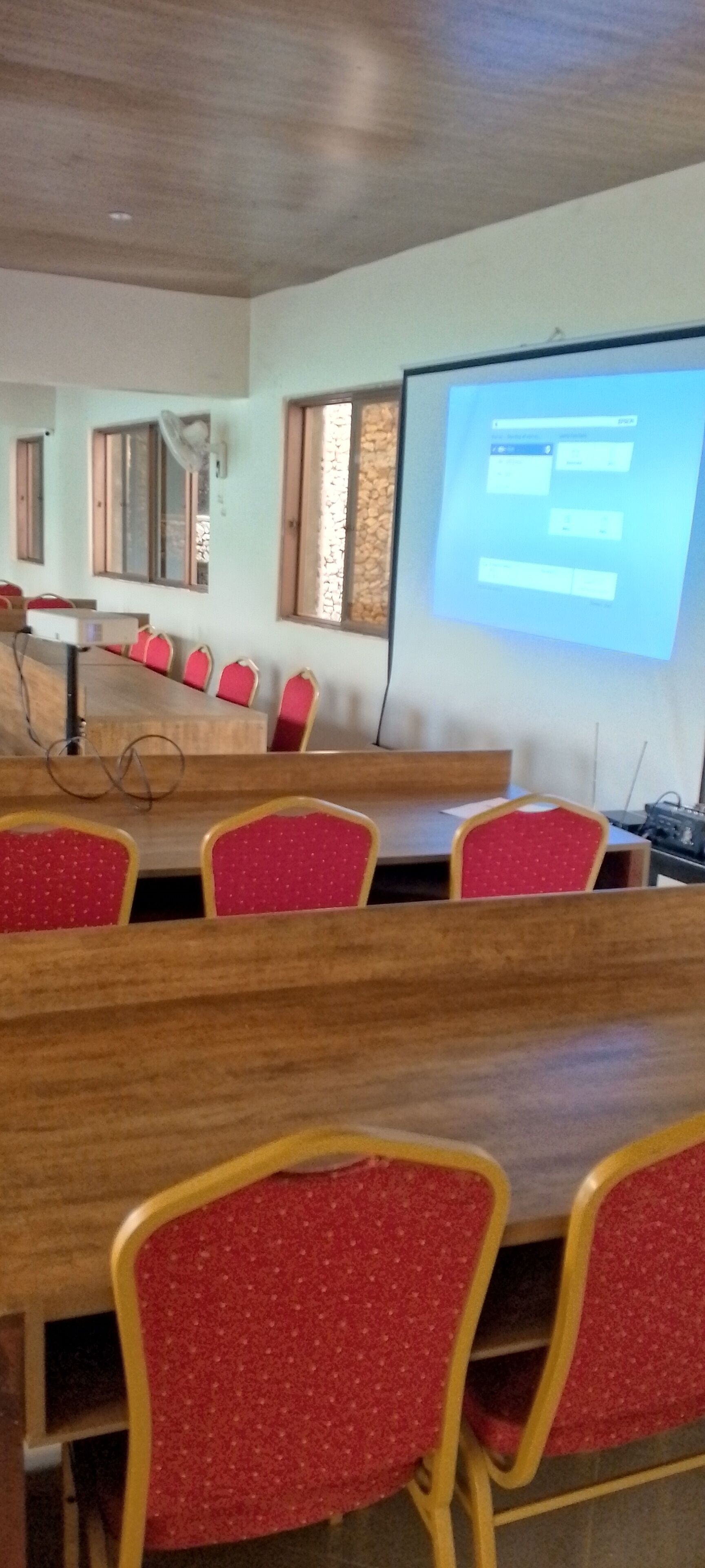Un salón de conferencias con una larga mesa y sillas rojas, una pantalla de proyector y ventanas con persianas.