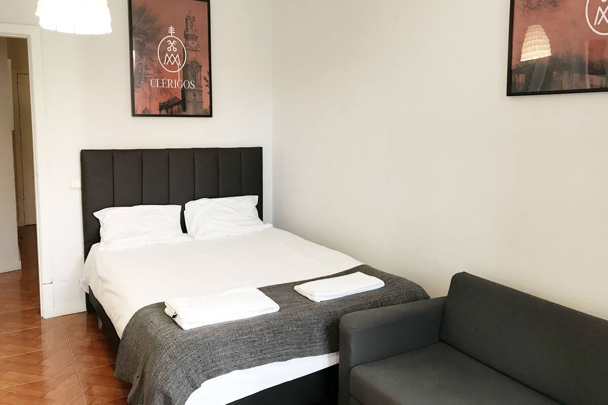 A neatly made bed with white linens, a dark headboard, and a grey sofa. There are two framed pictures on the wall, one of a building and another with a logo.