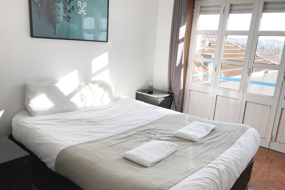 A neatly made bed with white linens and two towels, a small bedside table with a lamp, and a window with curtains.