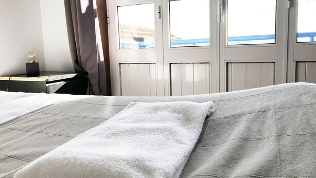 A bed with a white towel, a nightstand with a lamp, and a window with a view of the sea. - Clérigos Apartments (Porto)