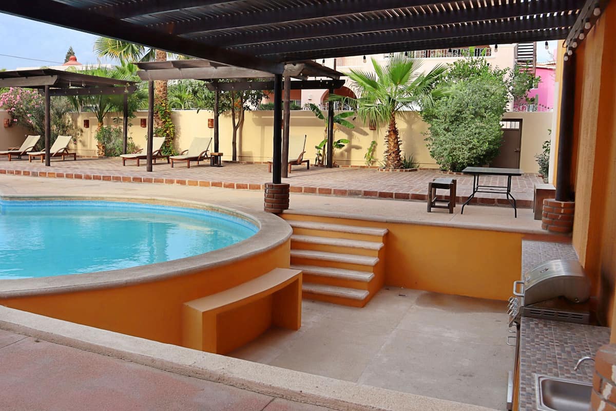 A swimming pool with a curved edge, a covered patio area with lounge chairs, and a barbecue grill.