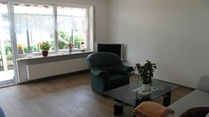 Un salón con sofá, una silla, una mesita de centro, televisor y ventana con plantas.