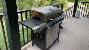 Een draagbare gasgrill op een houten terras met een hekwerk, omringd door begroeiing.