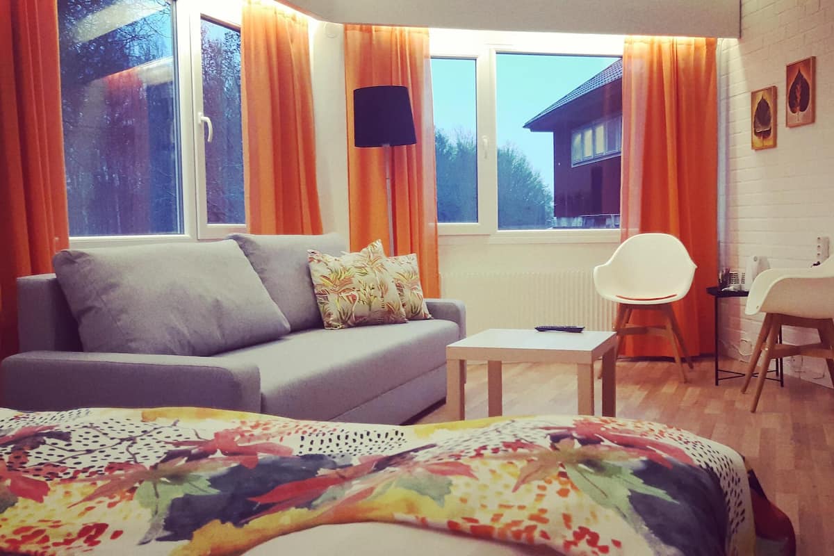A modern hotel room with a grey sofa, a small table, a white chair, and a bed with a colorful blanket.