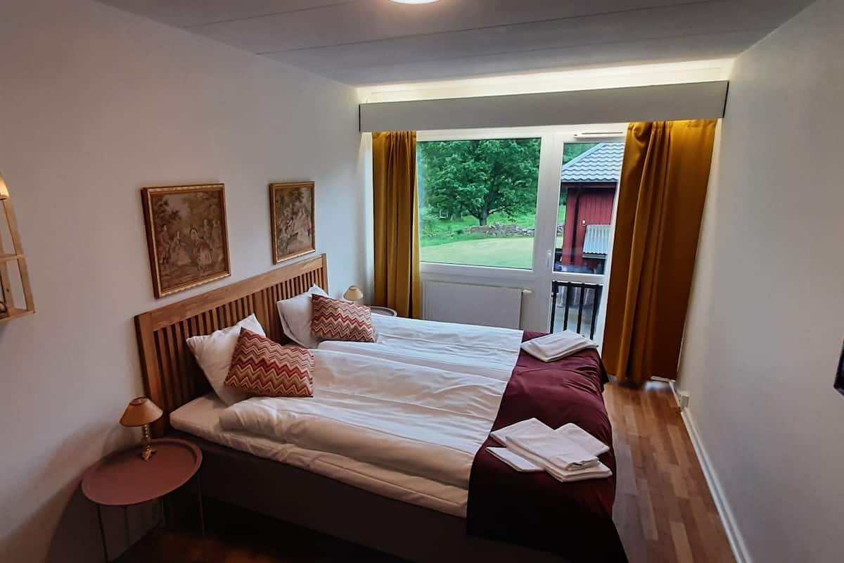 A hotel room with a bed, a bedside table with a lamp, a shelf with glassware, and a window with curtains.