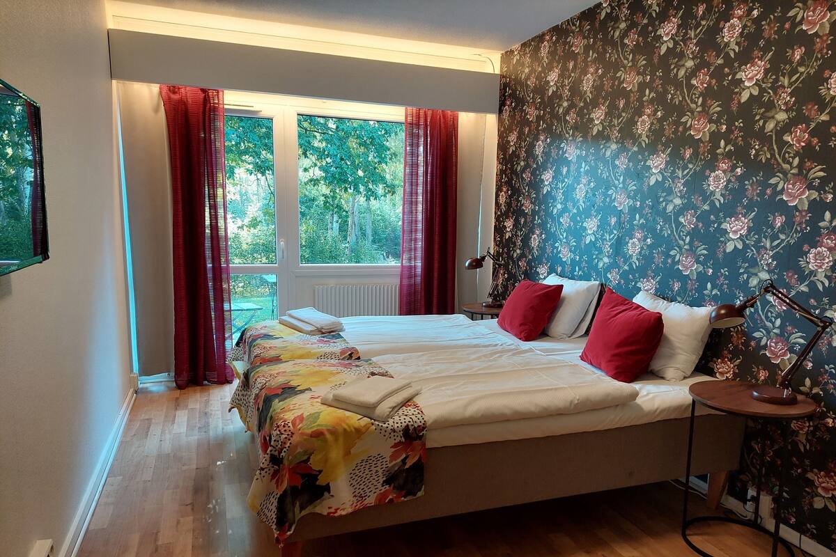 A bedroom with a large bed, red and white bedding, a floral wallpaper, and a view of greenery outside.