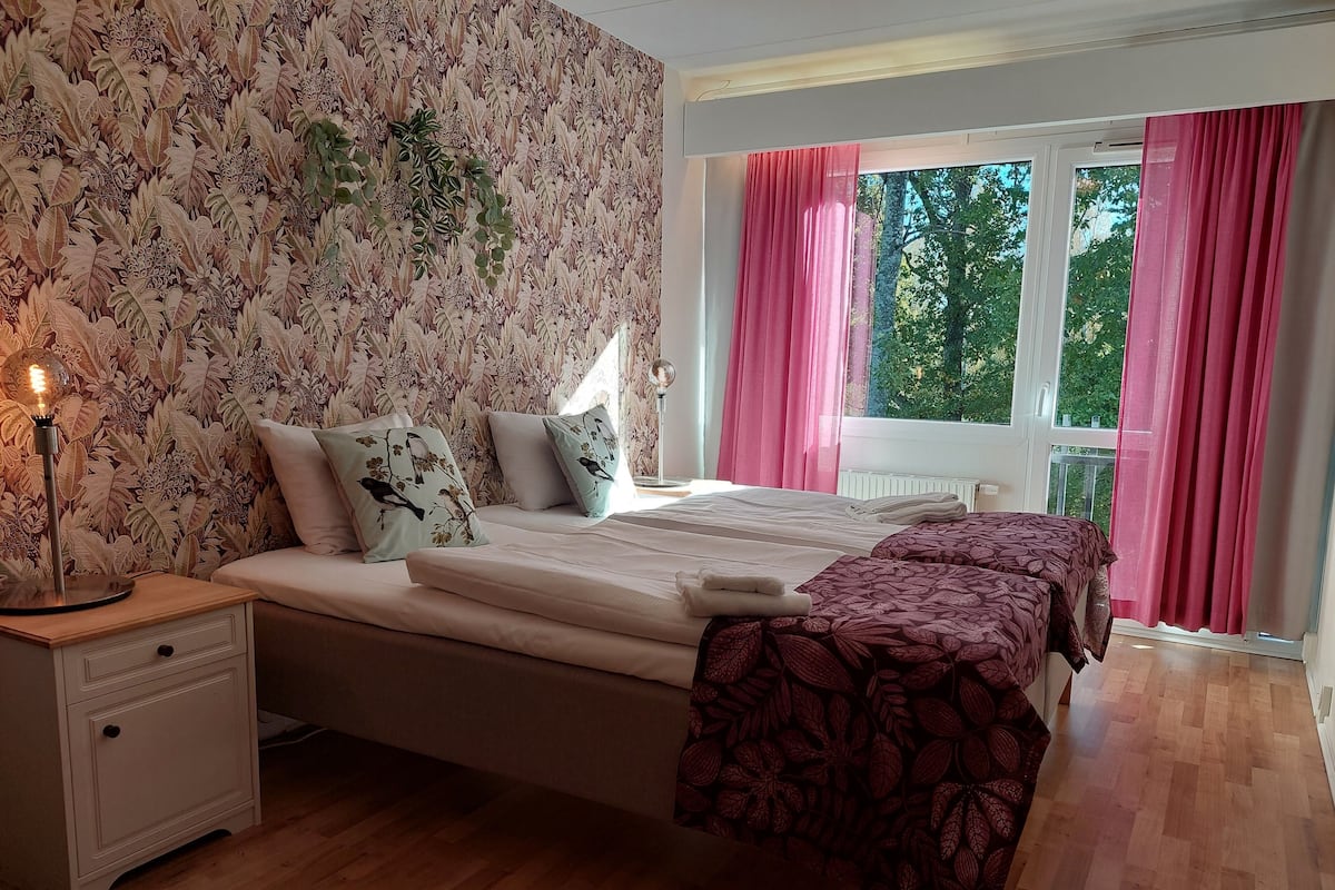 A bedroom with a floral wallpaper, a bed with pillows, a bedside lamp, and a window with curtains.