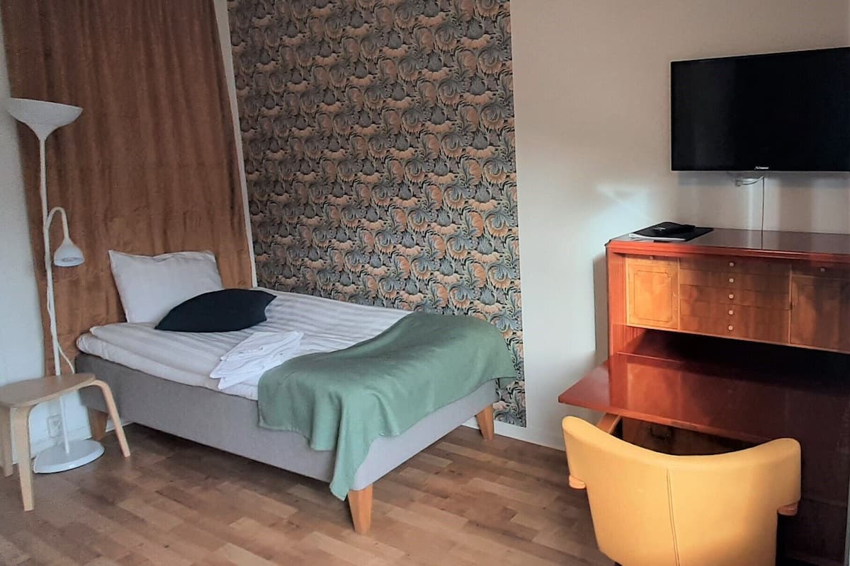 A room with a bed, a desk, a chair, a television, and a patterned wallpaper.