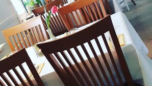 A dining area with wooden chairs and tables, a potted plant, and a view of the kitchen in the background.