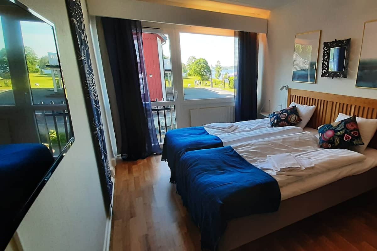 A hotel room with a bed, two blue bedsheets, a wooden headboard, a window with a view of a green landscape, and a door leading outside.