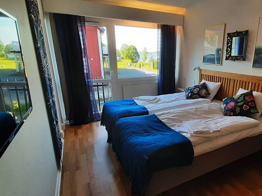Habitación de hotel con cama, dos sábanas azules, cabecera de madera, ventana con vistas a un paisaje verde y puerta que da al exterior.