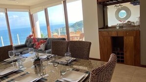 Ein Essbereich mit einem Tisch für sechs Personen, großen Fenstern mit Blick und einer Küchenzeile mit Holzschränken.