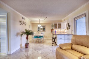 A spacious living area with a beige sofa, a dining table with chairs, a kitchen area with cabinets, and a potted plant.