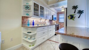 A kitchen with white cabinets, a granite countertop, and a backsplash. There are shelves with decorative items and a microwave above the counter. - A Little Slice of Paradise !! (Bonita Springs)
