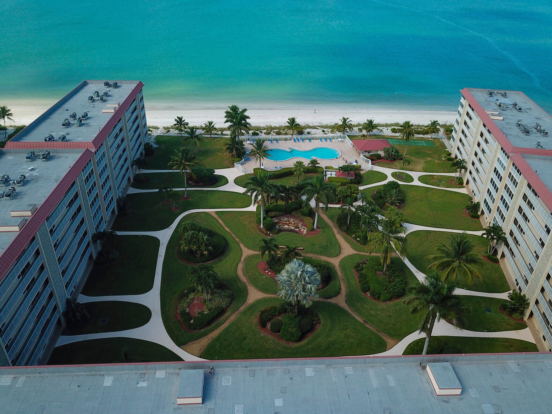 An aerial view of a hotel with a swimming pool surrounded by landscaped gardens and palm trees, overlooking a beach.