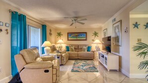 A living room with a beige sectional sofa, a ceiling fan, a TV, and a decorative wall with palm trees and stars. - A Little Slice of Paradise !! (Bonita Springs)