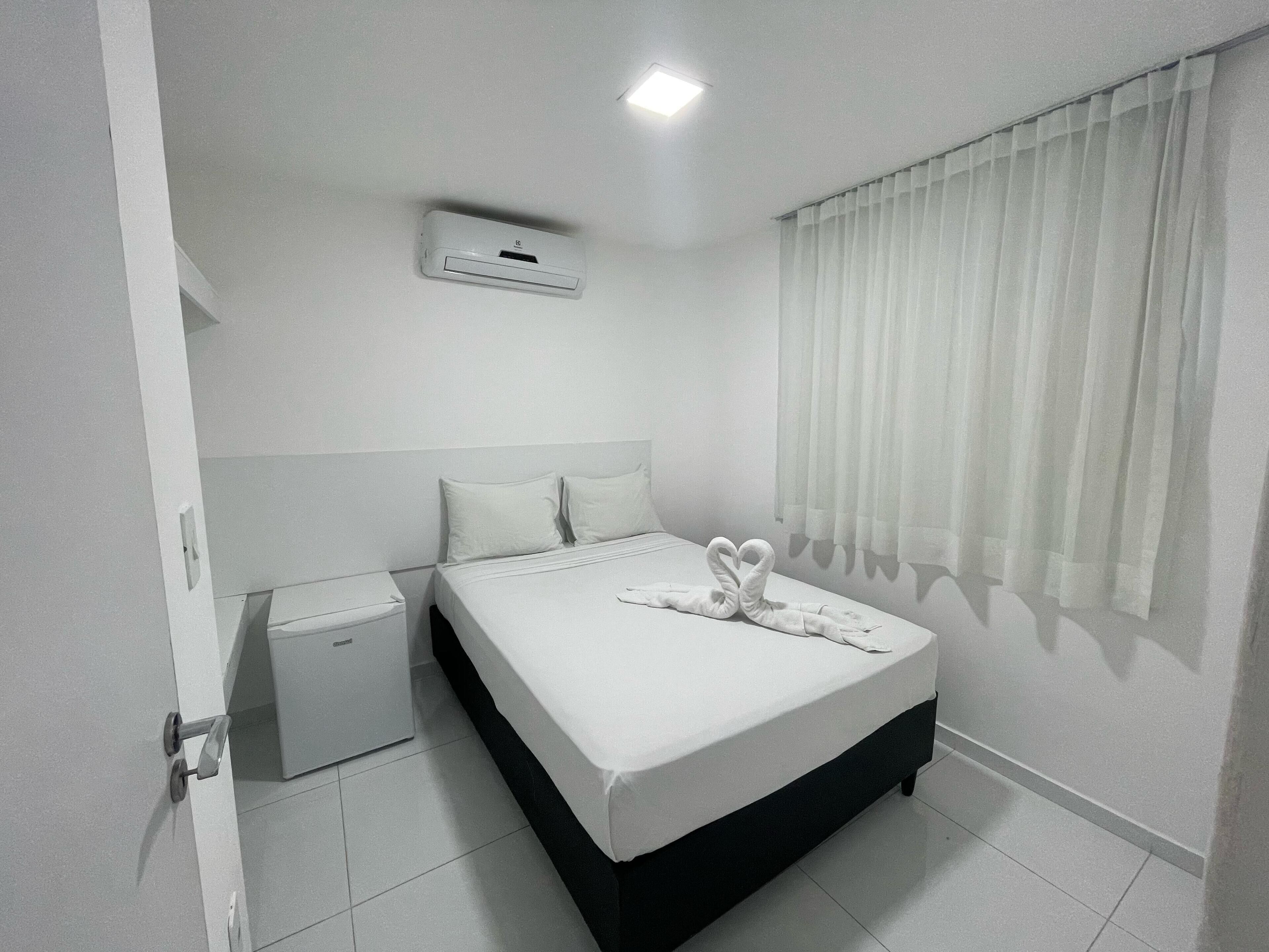 A hotel room with a bed, a bedside table, a small cabinet, and a window with curtains.