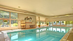 A large indoor swimming pool with clear views of a forest through large windows, a seating area with tables and chairs, and a small shelf with decorative items. - Spacious Remodel, 4br, Minutes To Conf Center & Slopes! 4 Bedroom Condo by RedAwning (Keystone)