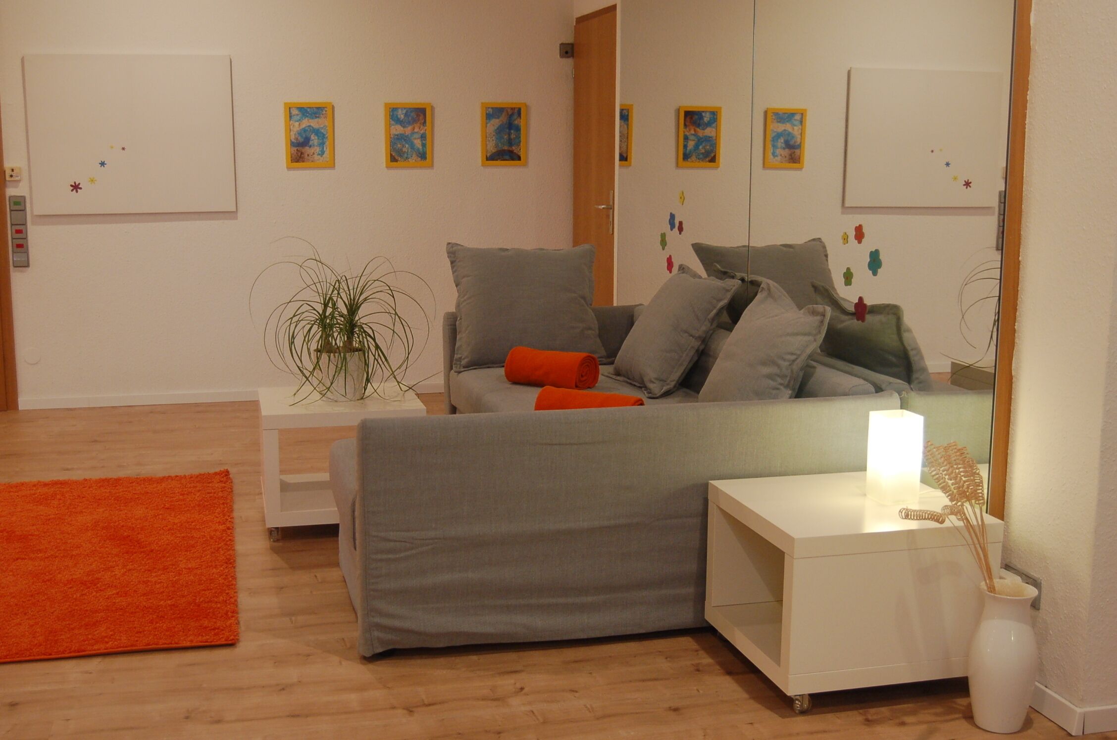 Een moderne woonkamer met een grijze bank, oranje kussens, een witte bijzettafel met een lamp en een kleine plant.