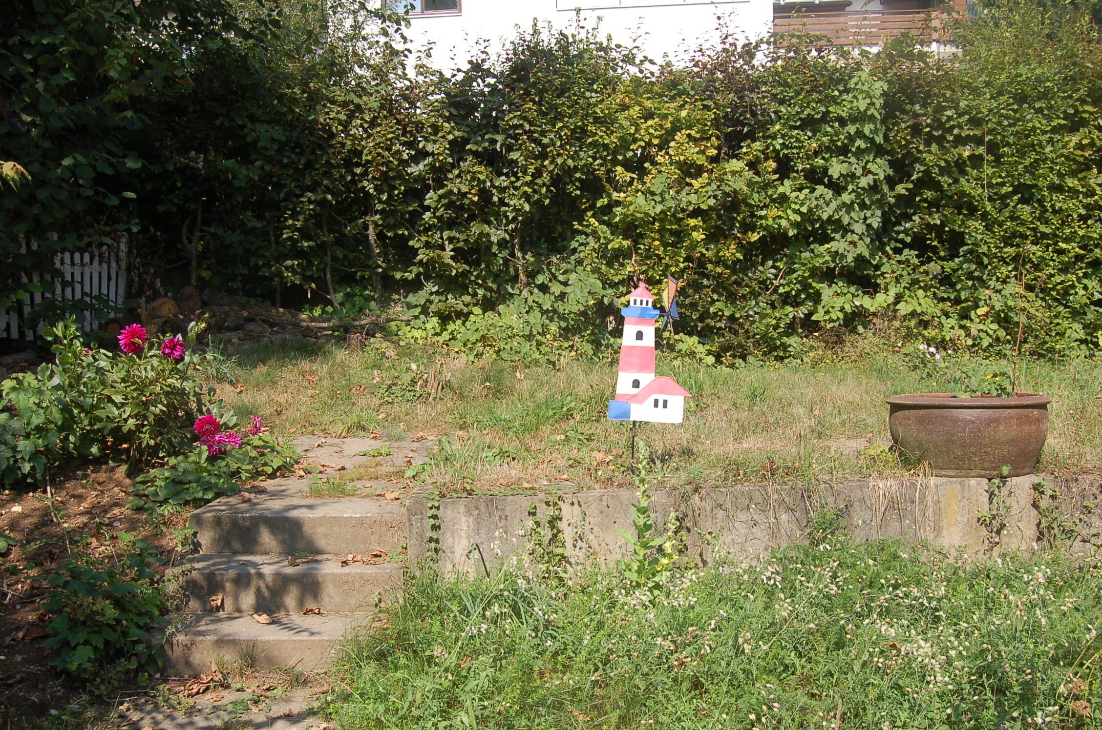 Une petite maquette de phare colorée, placée dans un jardin avec un mur en béton et un pot de fleurs à proximité.
