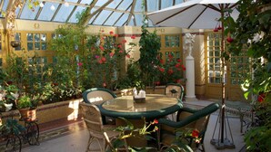 Un invernadero con techo de cristal, rodeado de macetas con plantas y flores, con una mesa redonda y sillas.