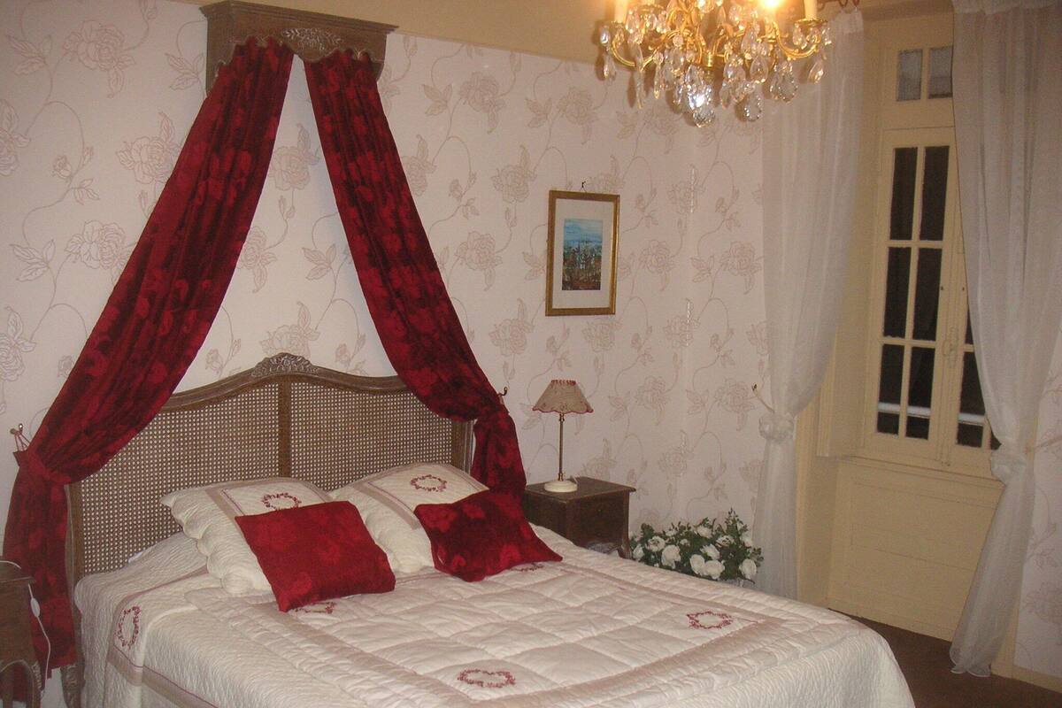 Une chambre à coucher avec un lit à baldaquin, un lustre, une table de chevet avec une lampe et une fenêtre avec des rideaux.