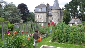 Un jardín con bancales elevados y diversas plantas, un edificio de piedra con una torre y una valla de madera.