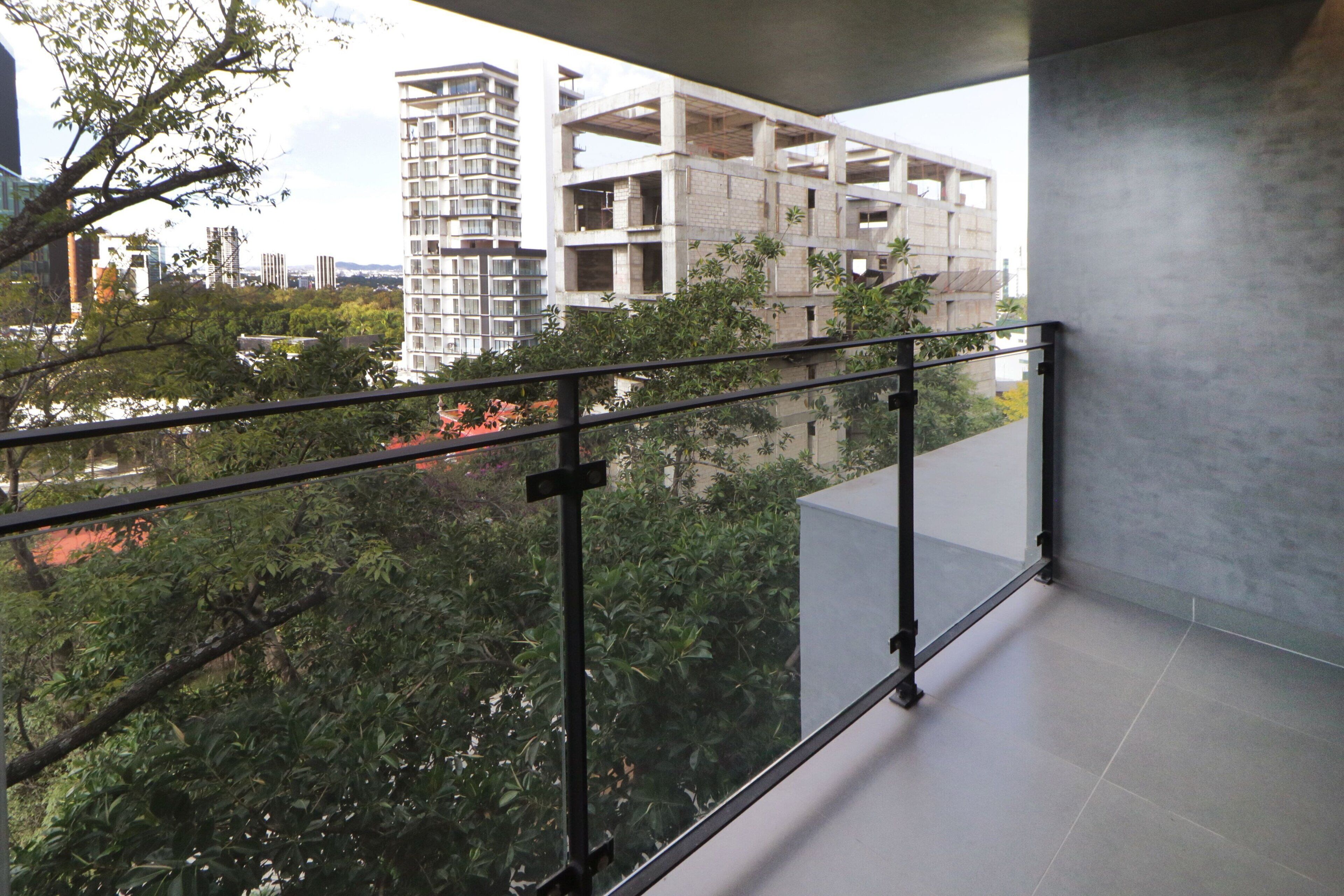 Balcón con baranda de metal que da a una obra en construcción y follaje circundante.