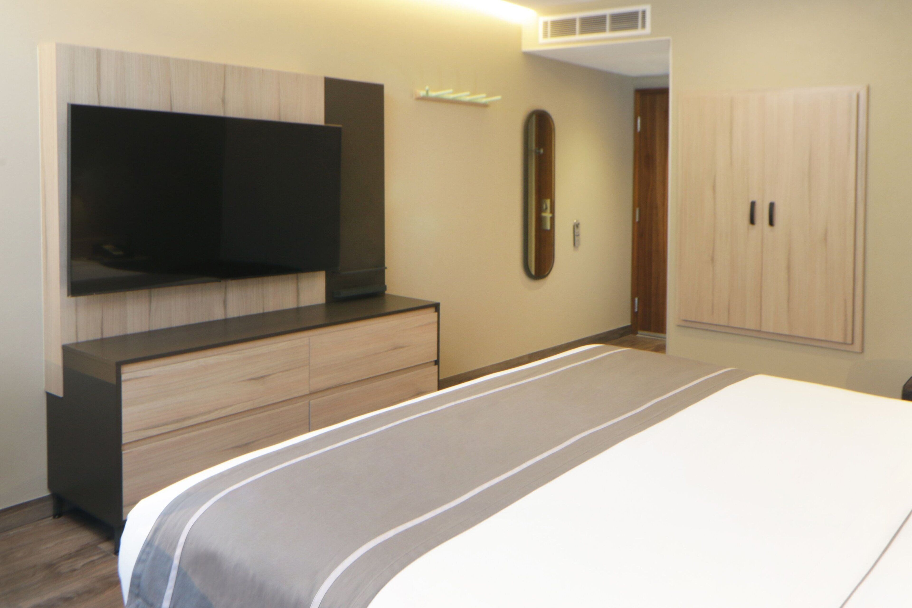 A bedroom with a bed, a TV mounted on the wall, a dresser, a mirror, and a wardrobe.