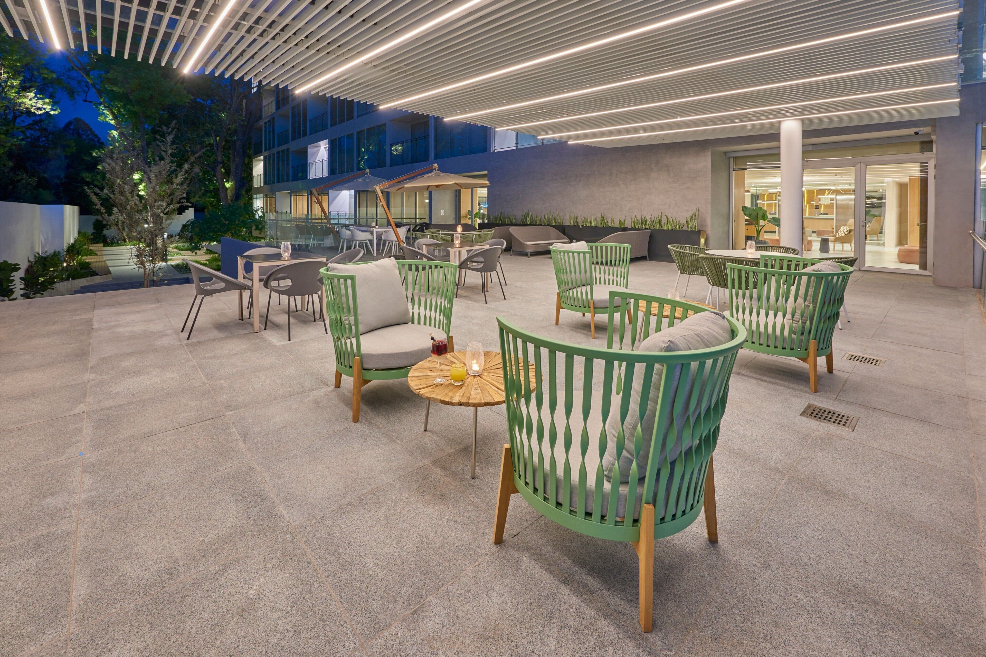 Un área de estar al aire libre con sillas verdes y mesas redondas, rodeada por un edificio moderno con amplias ventanas y un paseo techado.