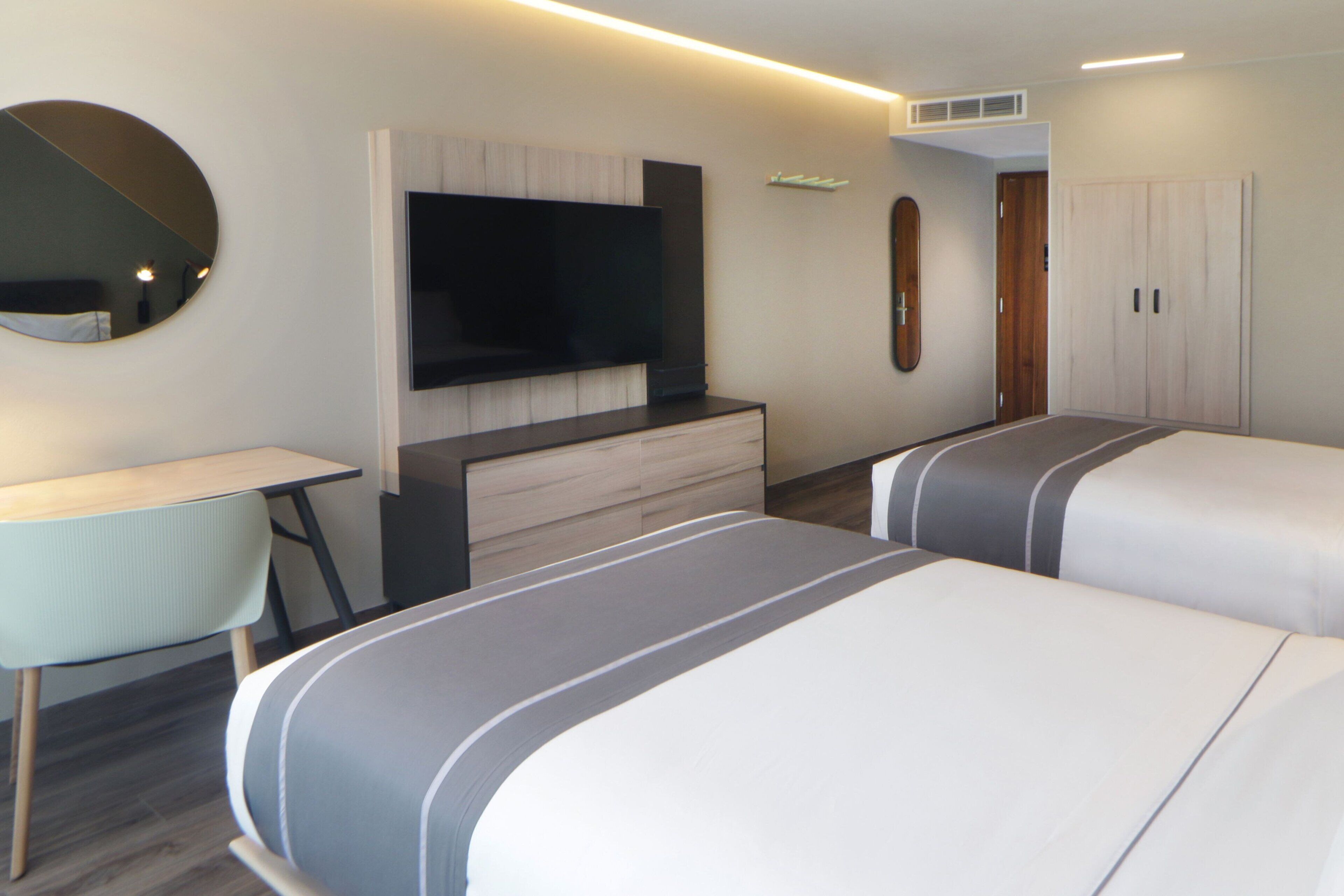 A modern hotel room with a flat-screen TV mounted on the wall, a desk with a chair, and two beds with striped bedding.