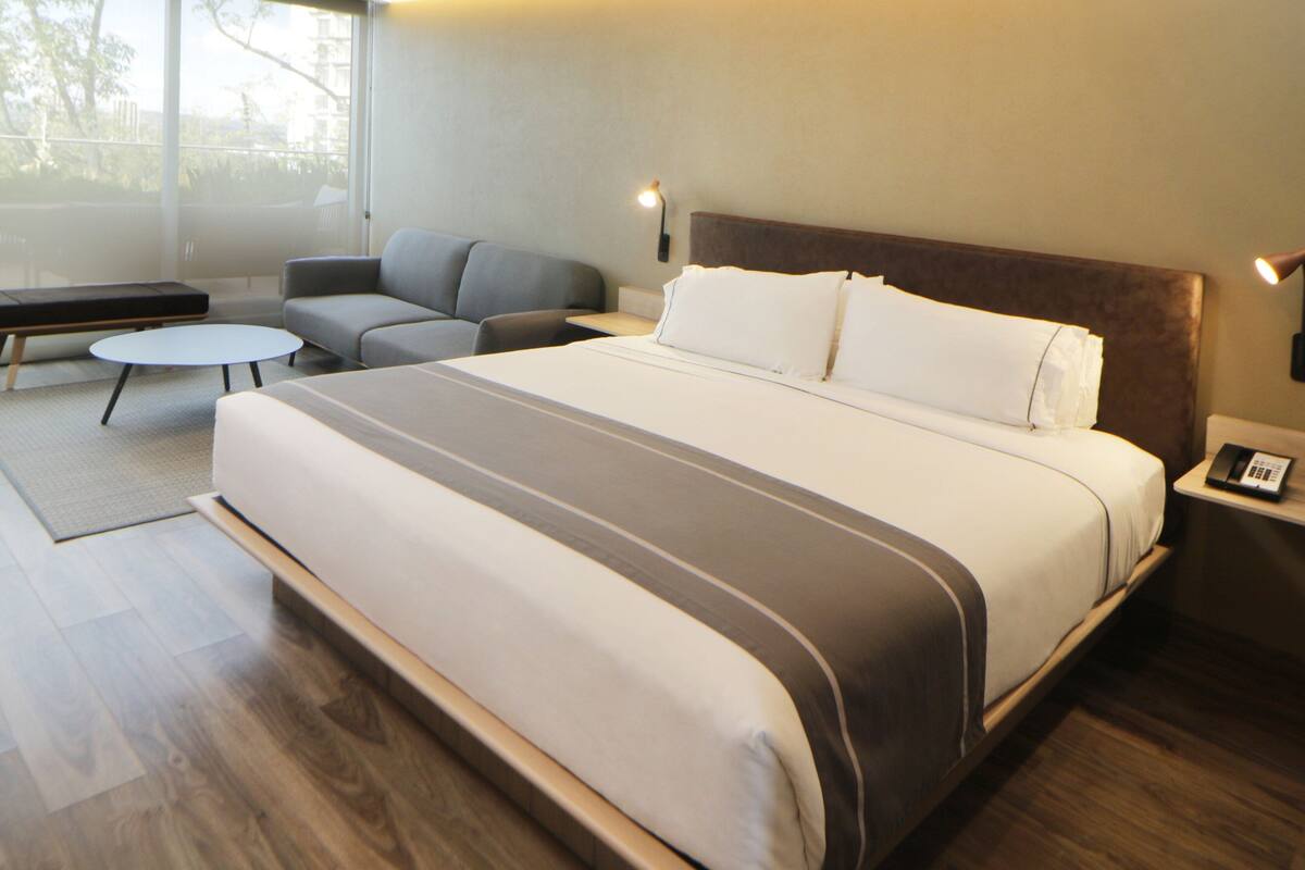 Una camera d'albergo moderna con un letto grande, un divano, un tavolino rotondo e un comodino con una lampada.