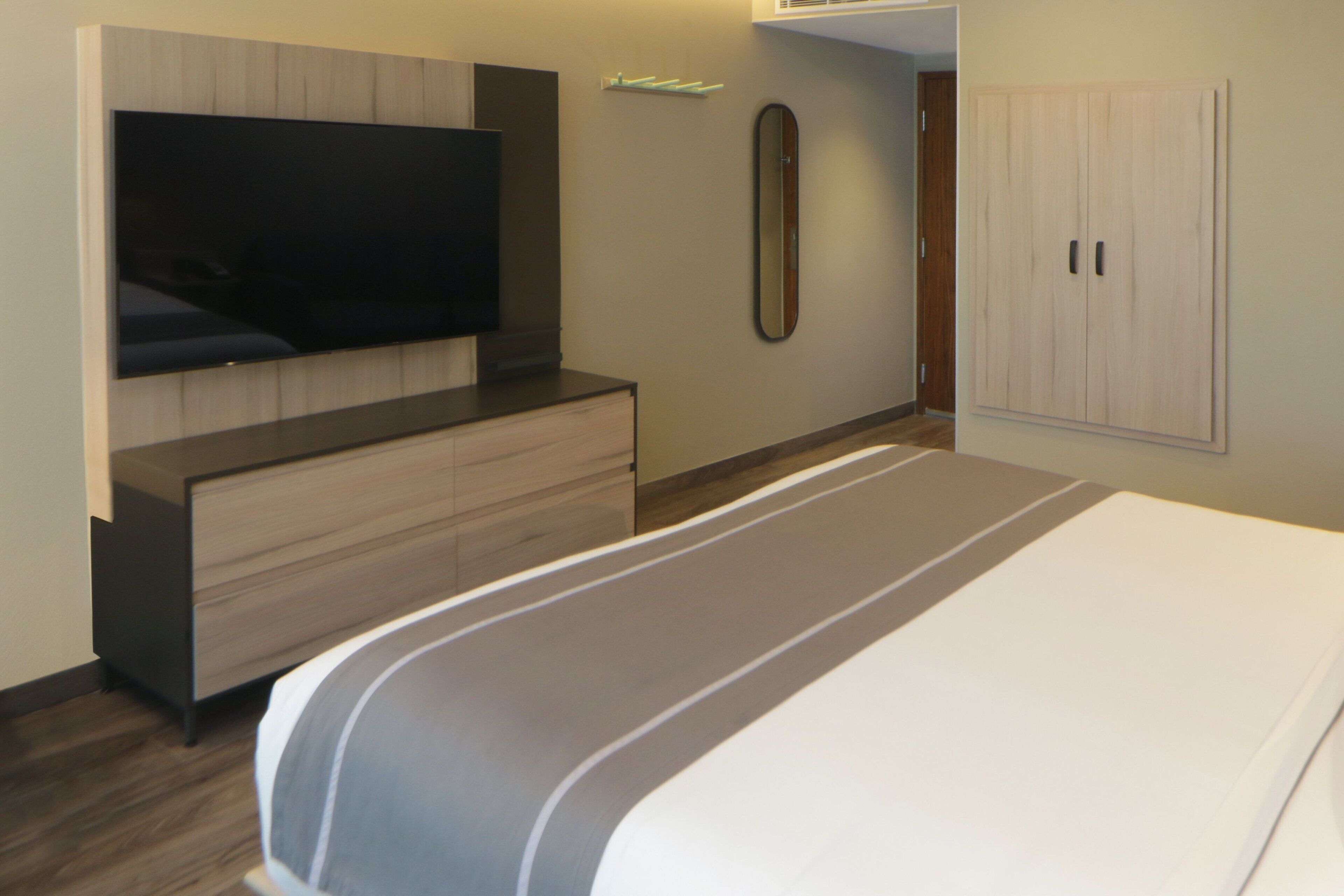 A modern bedroom with a large bed, a flat-screen TV mounted on the wall, a dresser, and a wardrobe.