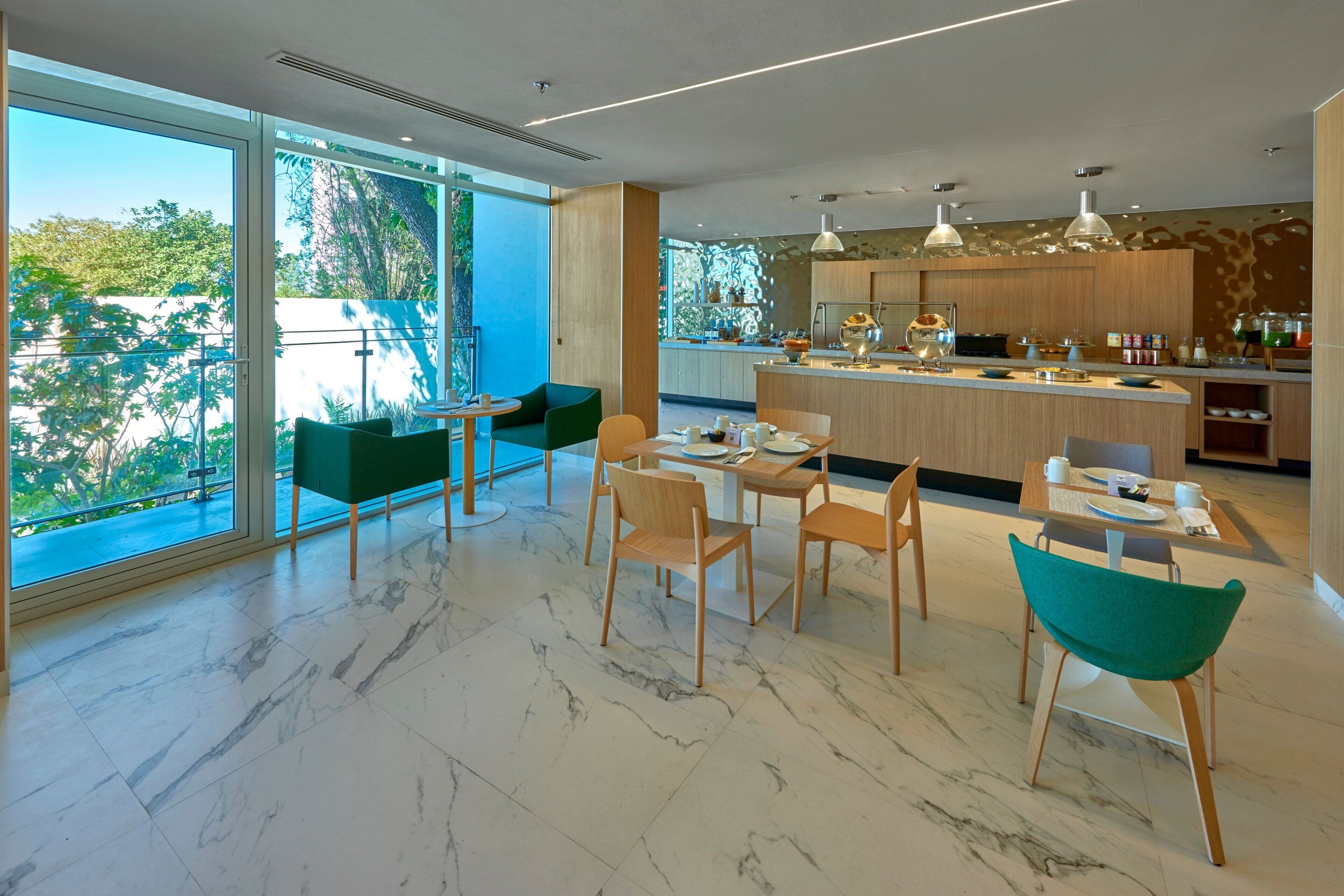 A modern kitchen with a marble floor, wooden cabinets, and a large island. There are two dining areas with green chairs and a table.