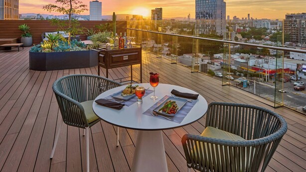 A rooftop terrace with a table set for dining, surrounded by wicker chairs, and a cityscape view at sunset. - City Express Plus by Marriott Guadalajara Providencia (Guadalajara)