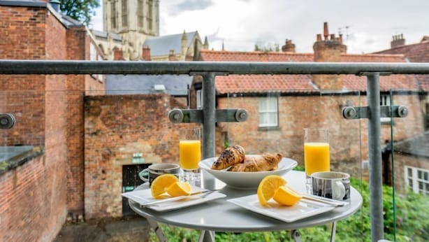 Un plateau de petit-déjeuner comprenant des croissants, du jus d’orange et une tasse, disposé sur un balcon donnant sur un bâtiment historique aux murs de briques et doté d’une tour.