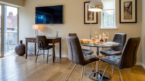 Un espace repas moderne avec une table en verre, des chaises à revêtement gris, un téléviseur à écran plat et des œuvres d’art encadrées accrochées aux murs.