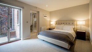 Une chambre moderne avec un grand lit, deux lampes de chevet, une cabine de douche en verre et une vue sur un bâtiment en briques, visible à travers une porte-fenêtre coulissante.