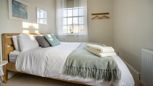 A neatly made bed with white linens and a grey throw blanket, a bedside table with a lamp, and a window with sheer curtains. - Riverside City Loft Apartment (York)