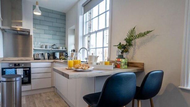 A modern kitchen with white cabinets, a stainless steel oven, a sink, and a window with a view. There are two blue chairs and a plant on the counter. - Riverside City Loft Apartment (York)