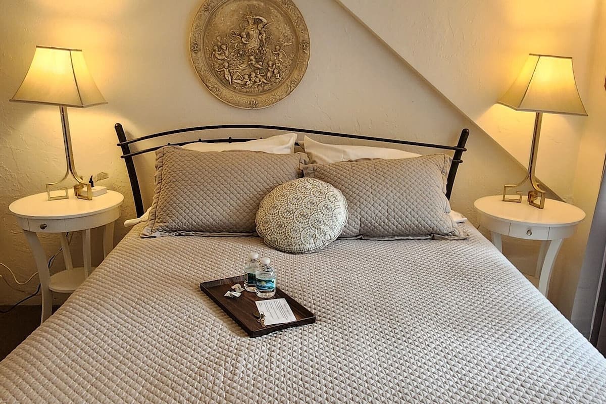 A neatly made bed with a quilted cover, two bedside tables with lamps, a decorative plate on the wall, and a tray with bottles and a tissue on the bed.