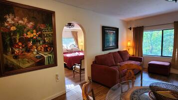 A living room with a sofa, a dining table with chairs, a painting, and a window with curtains.