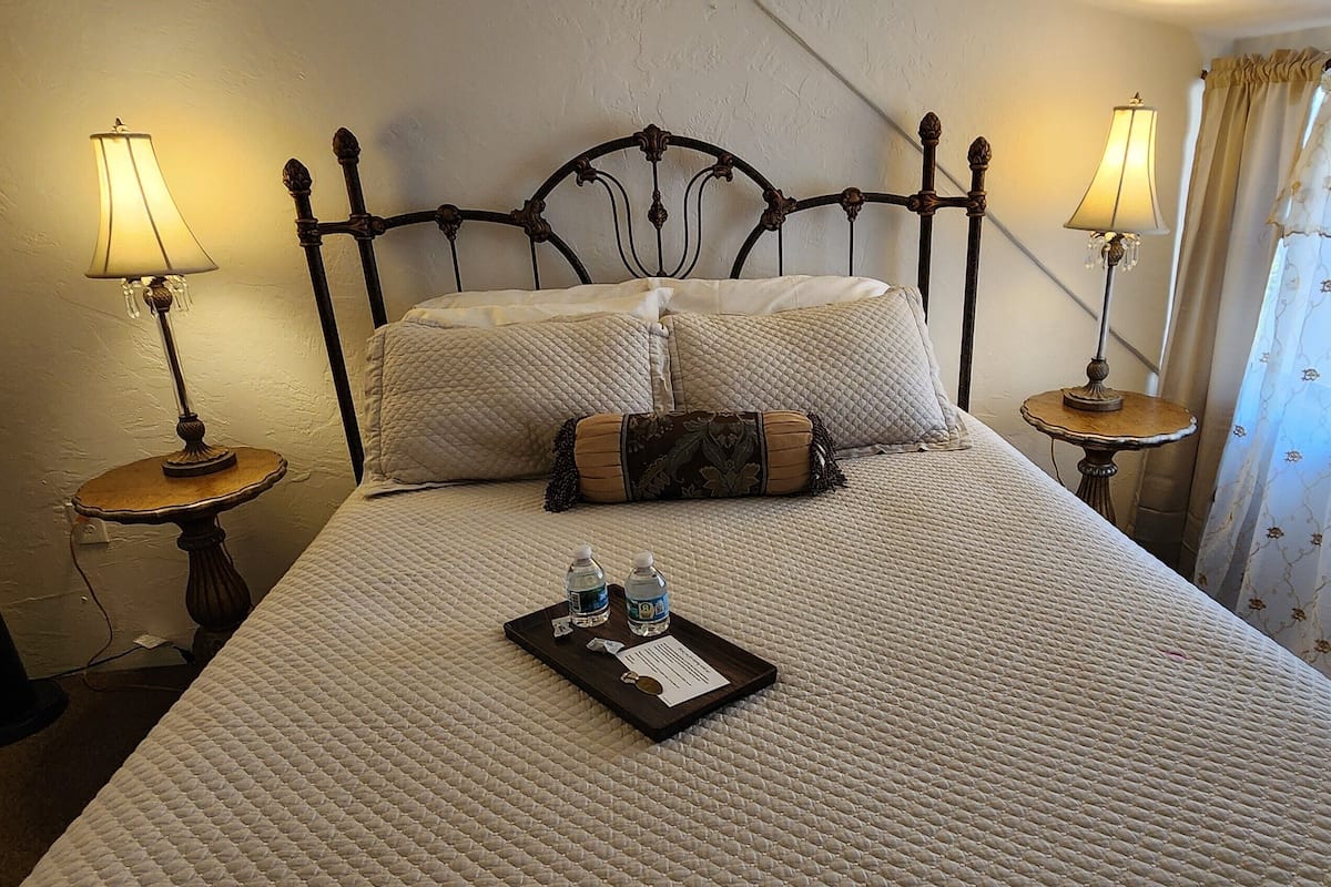 A neatly made bed with a quilted bedspread, two pillows, a tray with toiletries, and two bedside lamps on matching nightstands.