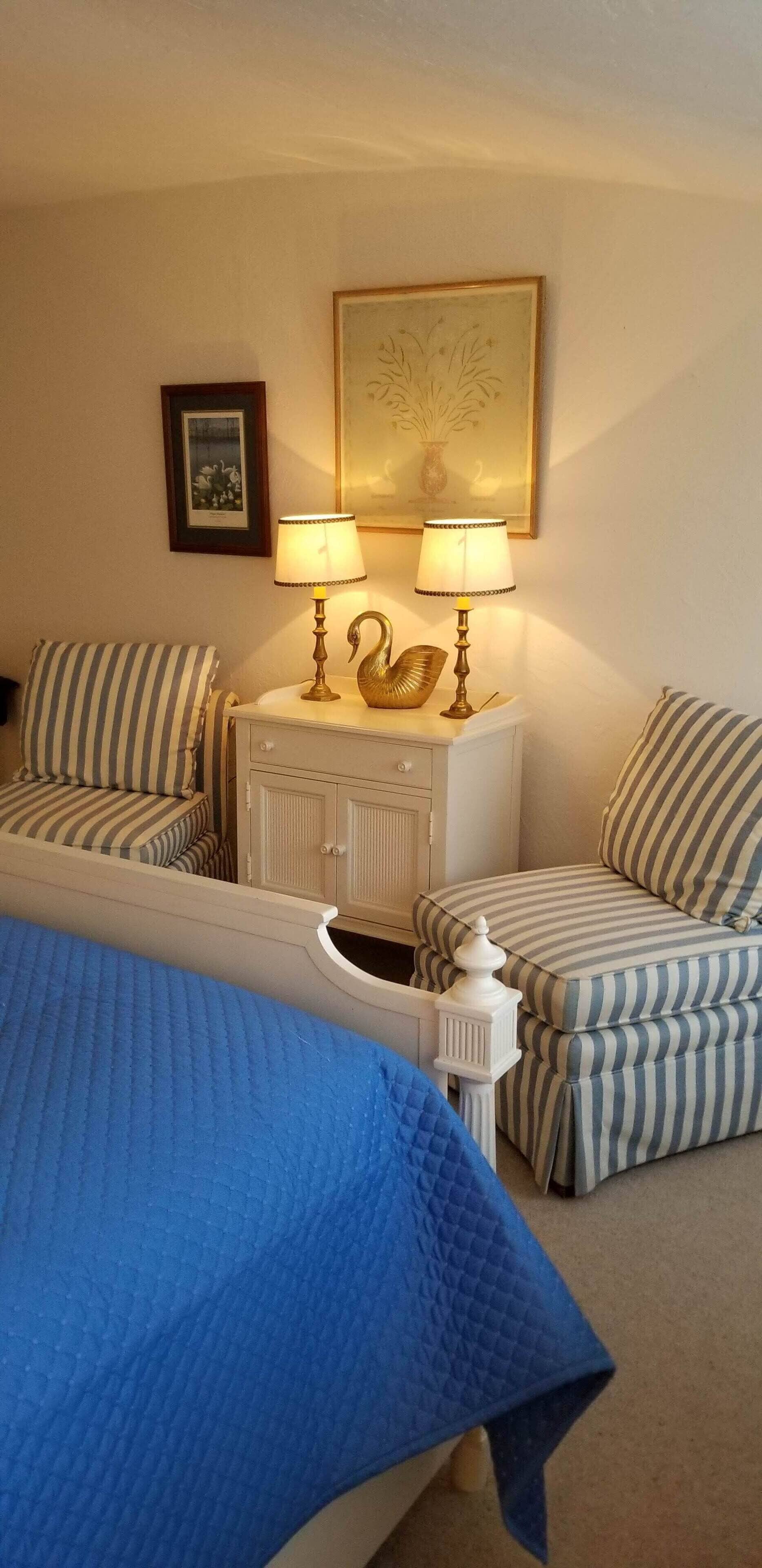 A hotel room with a bed, two bedside tables with lamps, striped upholstered chairs, a small cabinet, and a framed picture on the wall.