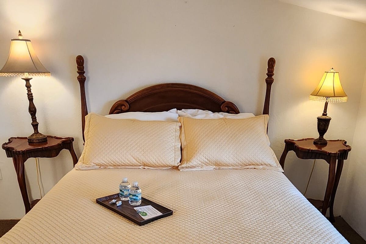 A neatly made bed with a quilted bedspread, two pillows, and a tray with water bottles and a note on the bed.